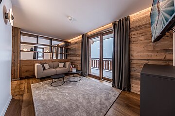A cozy, modern living room featuring warm wood walls, a beige sofa, and a large rug, with a balcony door revealing a snowy outdoor view.