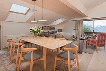 Modern chalet interior featuring a light wood dining table under skylights, leading to a living area with a scenic mountain view.