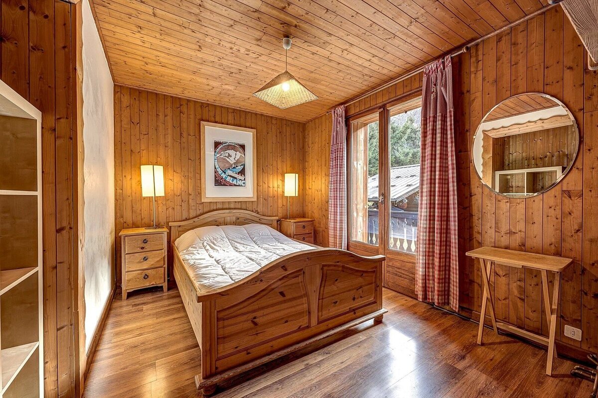 A cozy, wood-paneled bedroom with a rustic wooden bed, red plaid curtains, large window, round mirror, and a framed skiing poster.