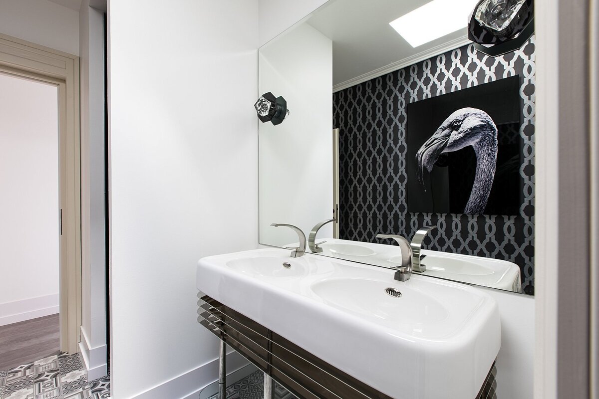 A stylish bathroom featuring a white double-sink vanity, a large mirror, and patterned floor tiles. The mirror reflects black wallpaper and a flamingo artwork.