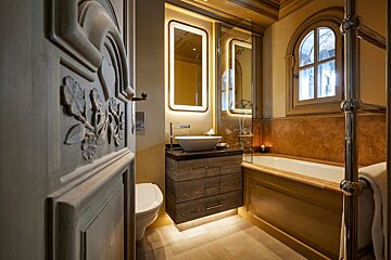 A luxurious, warm-lit bathroom with a carved wooden door, illuminated mirror, vessel sink, bathtub, and arched window.