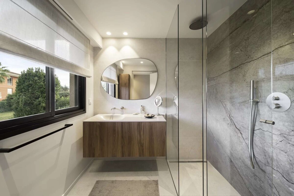 A modern bathroom features a floating wood vanity, unique mirror, glass shower with stone accents, and a window overlooking lush greenery.