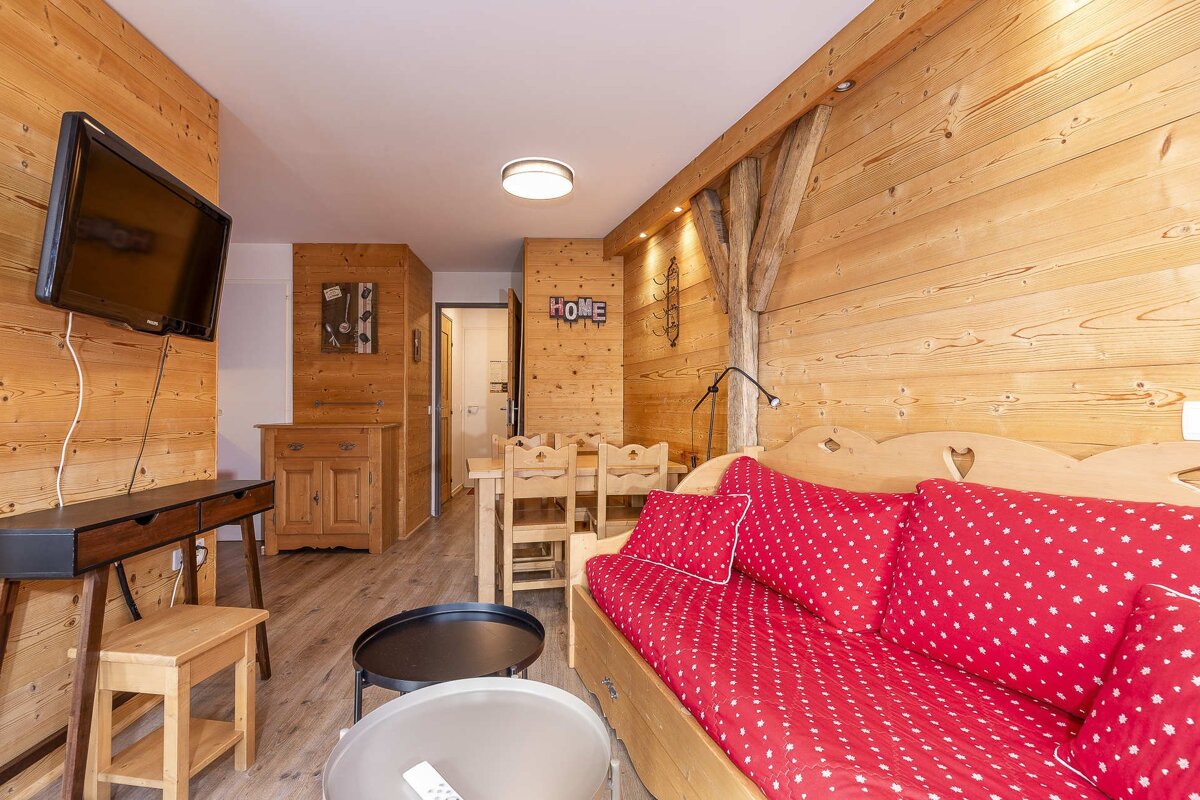 A warm, wood-paneled living area featuring a TV, dining table, and a red patterned daybed in a cozy, rustic, chalet-style setting.