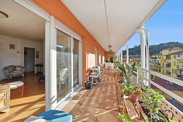 A long, sunlit balcony with orange walls, plants, and outdoor furniture overlooks yellow buildings and green hills. An indoor living space is visible.
