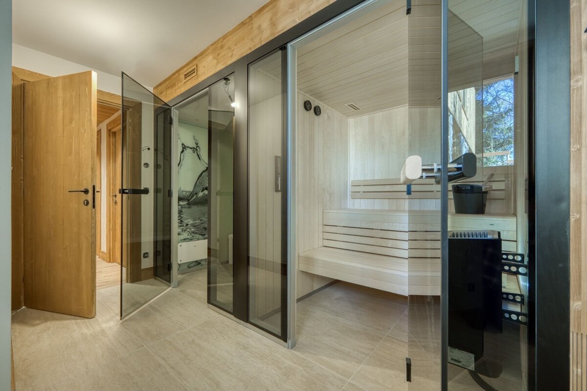 A modern room with a glass-enclosed wooden sauna, a shower stall featuring a water mural, and light wood doors in a tiled hallway.