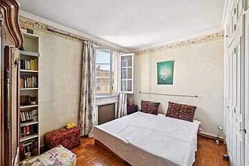 A compact bedroom with a bed, open window, filled bookshelf, and white wardrobe. Light walls with a floral border, wooden parquet floor, and a lotus painting.