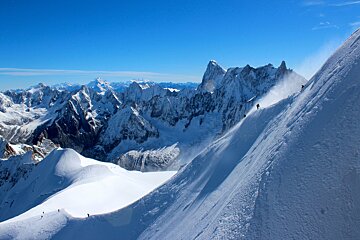 Skiers descend steep, powdery slopes amidst vast snow-capped mountains under a clear blue sky, kicking up snow as they go.