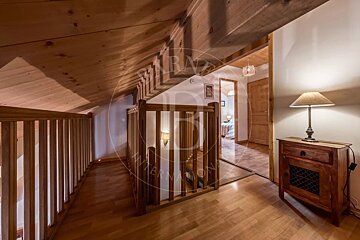 Rustic wooden interior with a sloping ceiling, featuring a landing area, a wooden balustrade, and a lamp on a cabinet. A doorway leads to another room.