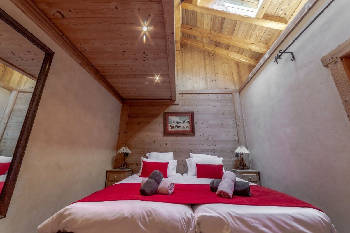 A cozy, wood-lined attic bedroom featuring twin beds with red throws, bedside lamps, a skylight, and rustic wall art.
