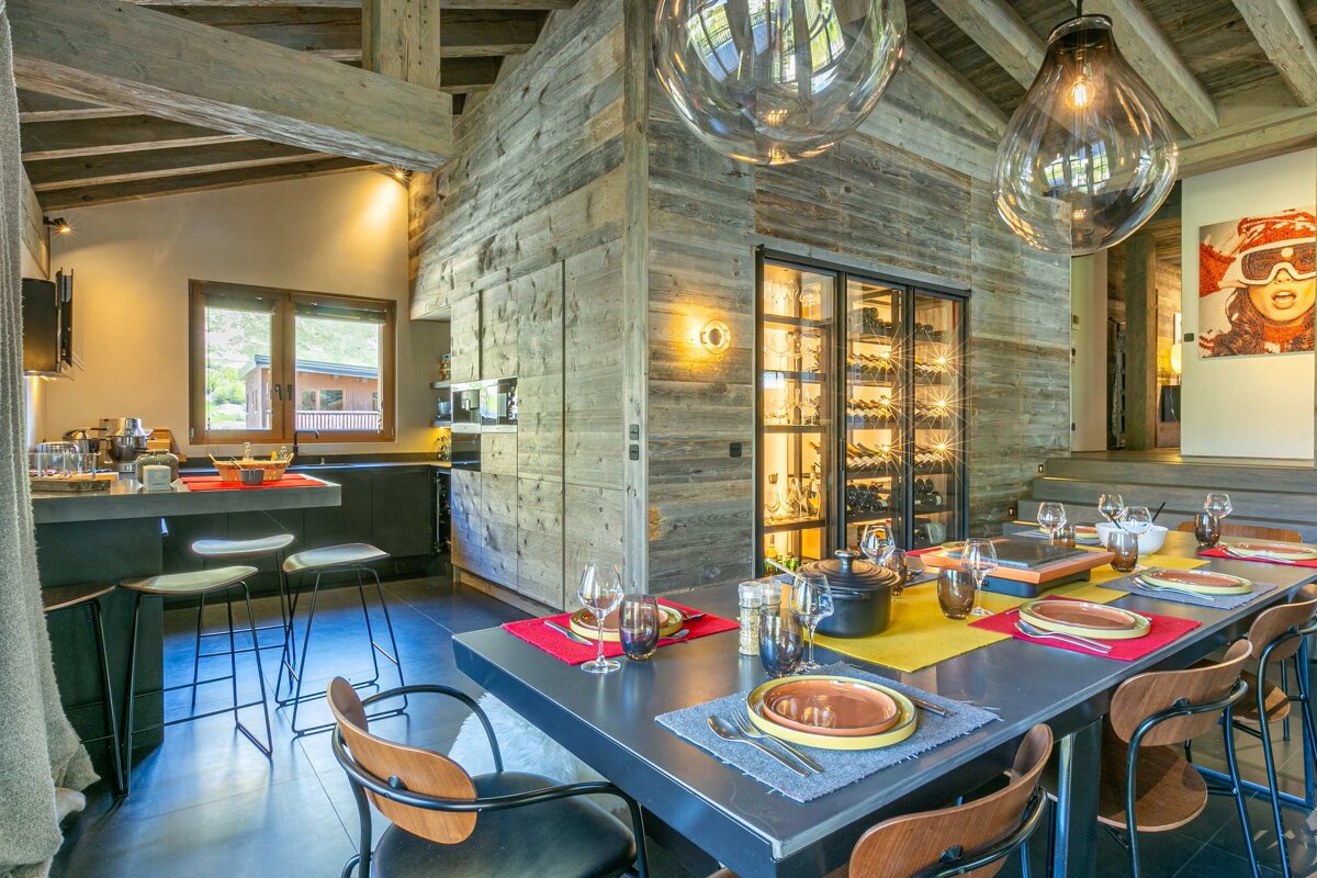 Rustic-chic chalet dining and kitchen area with exposed wood, a large set dining table, bar seating, and an illuminated wine cellar.