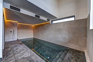 Modern indoor swimming pool area with grey tiles, ambient strip lighting, a shower, and steps leading into dark water.