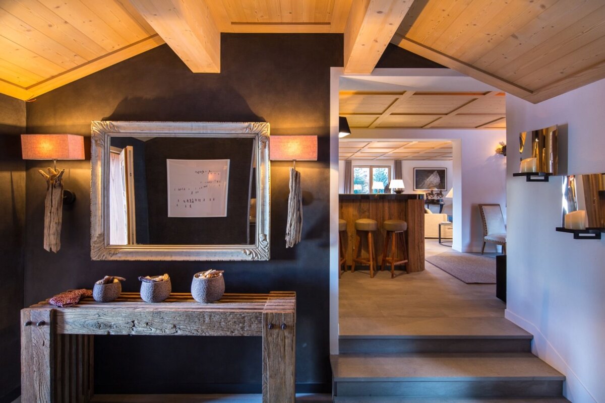 A rustic-chic interior hallway with dark walls, wooden beams, a large ornate mirror, and a wood console table, leading to a cozy bar area.