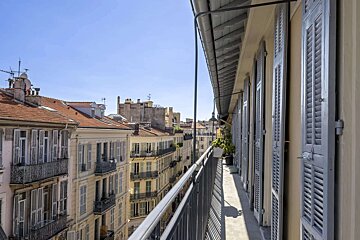 A long balcony with light-blue shutters overlooks a sunlit European city street filled with traditional buildings, terracotta roofs, and a clear sky.