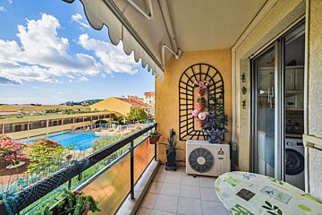 A decorated balcony with an awning overlooks a vibrant blue swimming pool and yellow buildings under a bright, cloudy sky.