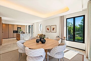 Modern open-plan dining and kitchen area with a round wooden table, upholstered chairs, marble island, and large windows looking out onto lush greenery.