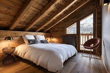A cozy, rustic chalet bedroom features warm wood panels & beams, a white bed, stylish lamps, and a balcony with a snowy mountain view.