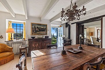 A rustic dining room features a long wooden table, ornate chandelier, and bright window/door leading to a garden with sea views, adjoining a cozy living area.