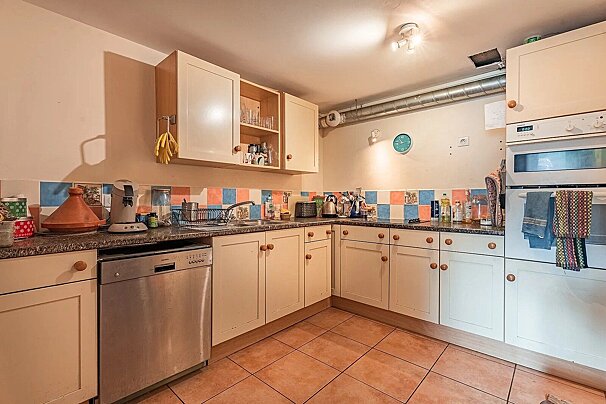 An L-shaped kitchen featuring cream-colored cabinets, a colorful tiled backsplash, a dishwasher, and terracotta tiled floor.