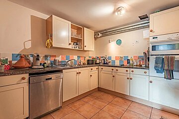 A cozy, cluttered kitchen with cream cabinets, blue and orange tiled backsplash, terracotta floor, stainless steel dishwasher, and built-in oven.