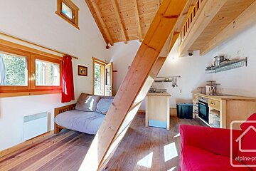 A bright, cozy loft studio featuring exposed wooden beams, a compact kitchen, a living area with a sofa, and red accents, bathed in sunlight.