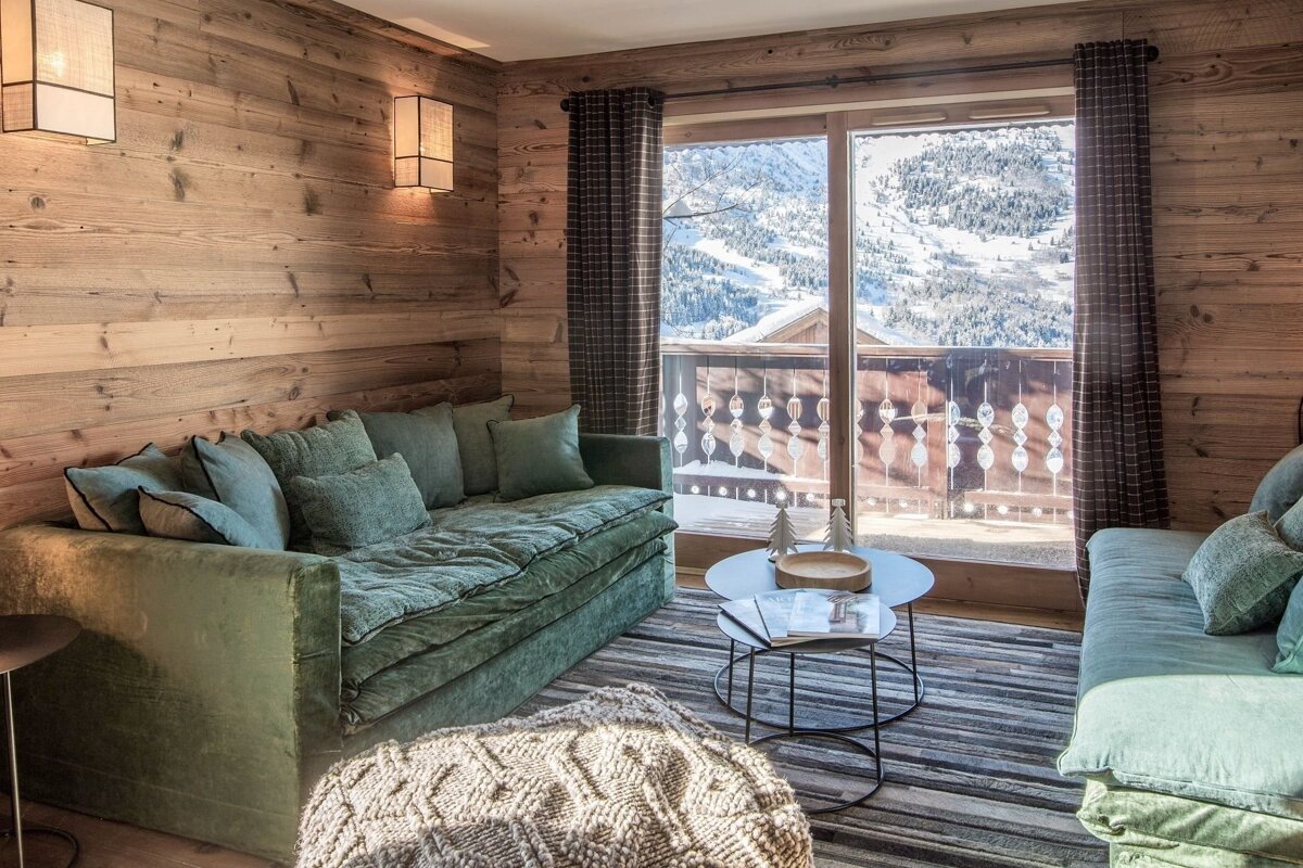 Cozy wood-paneled room with green sofas and warm lighting, featuring a stunning view of snowy mountains from a large balcony window.