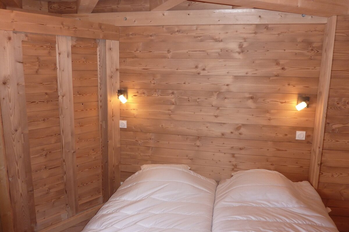 Cozy bedroom with light wood paneling on walls and ceiling. A bed with two white duvets is lit by warm wall-mounted lamps.