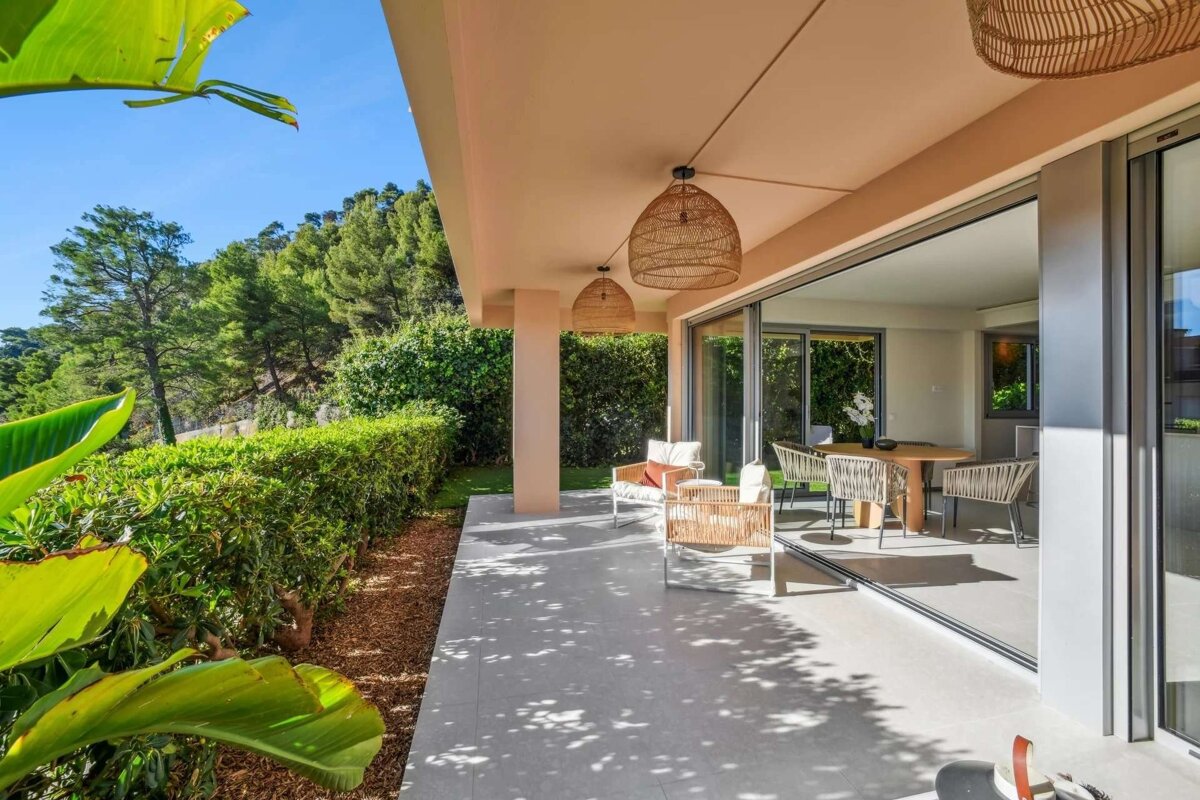 A chic outdoor patio with stylish rattan furniture, large glass doors, and wicker lights, overlooking a lush green hillside on a sunny day.