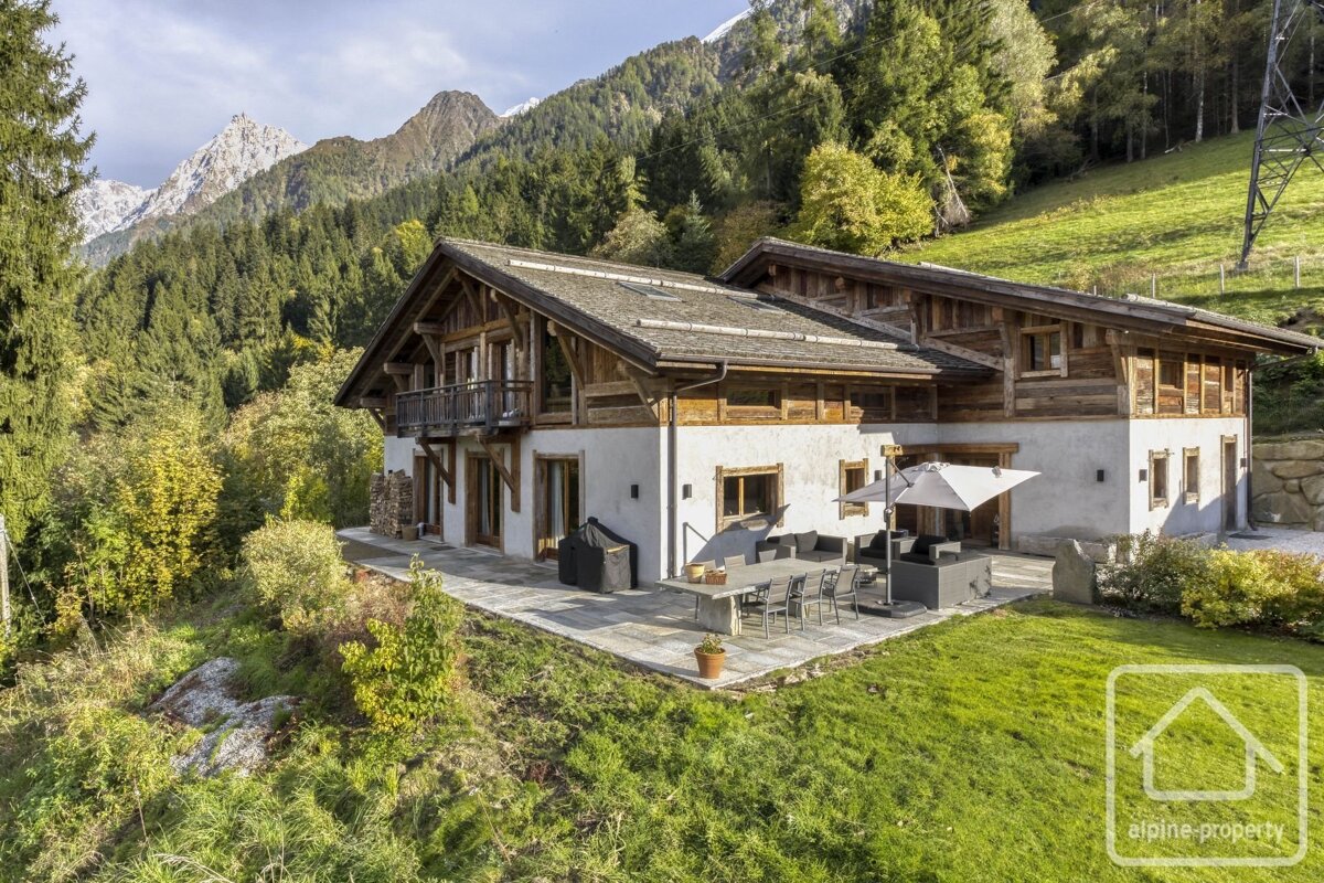 A large wooden chalet with a stone patio, nestled in a lush green valley with forested mountains and peaks in the background.