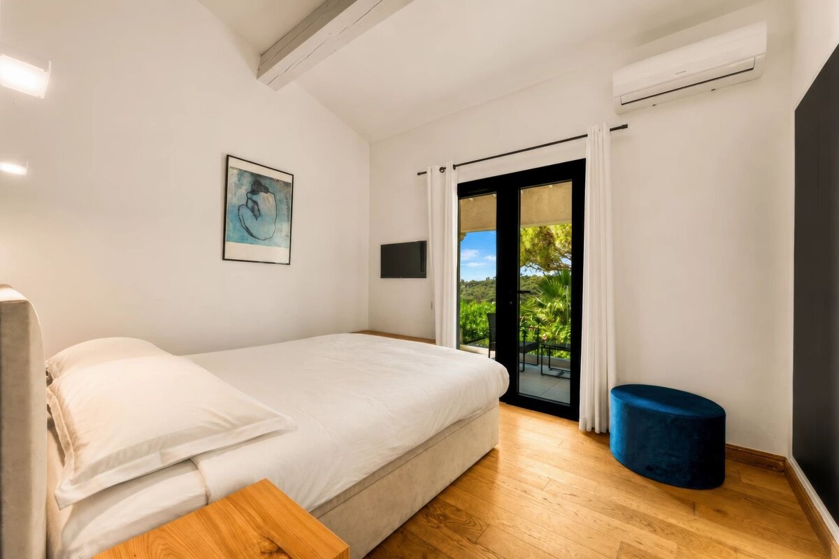 Modern bedroom with a white bed, light wood floors, a blue ottoman, and glass double doors opening onto a scenic balcony.