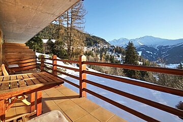 Wooden balcony with a table overlooking a snowy valley and mountains under a clear blue sky.