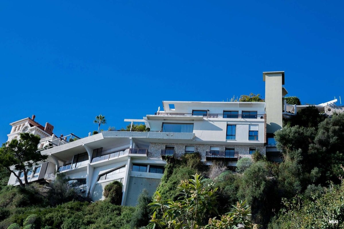 A modern multi-level white building and an ornate villa are nestled into a lush, green hillside under a clear blue sky.