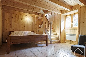 A cozy, rustic room with light wood paneling, ceiling beams, and a wooden staircase. Features a bed, tiled floor, and a bright window.