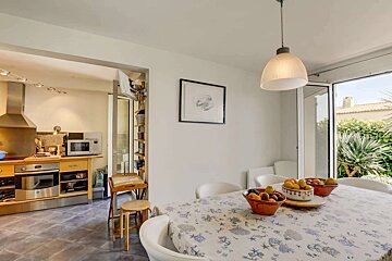 A bright dining area with a patterned tablecloth and fruit bowls, connected to a modern kitchen and offering a view of a lush garden through glass doors.