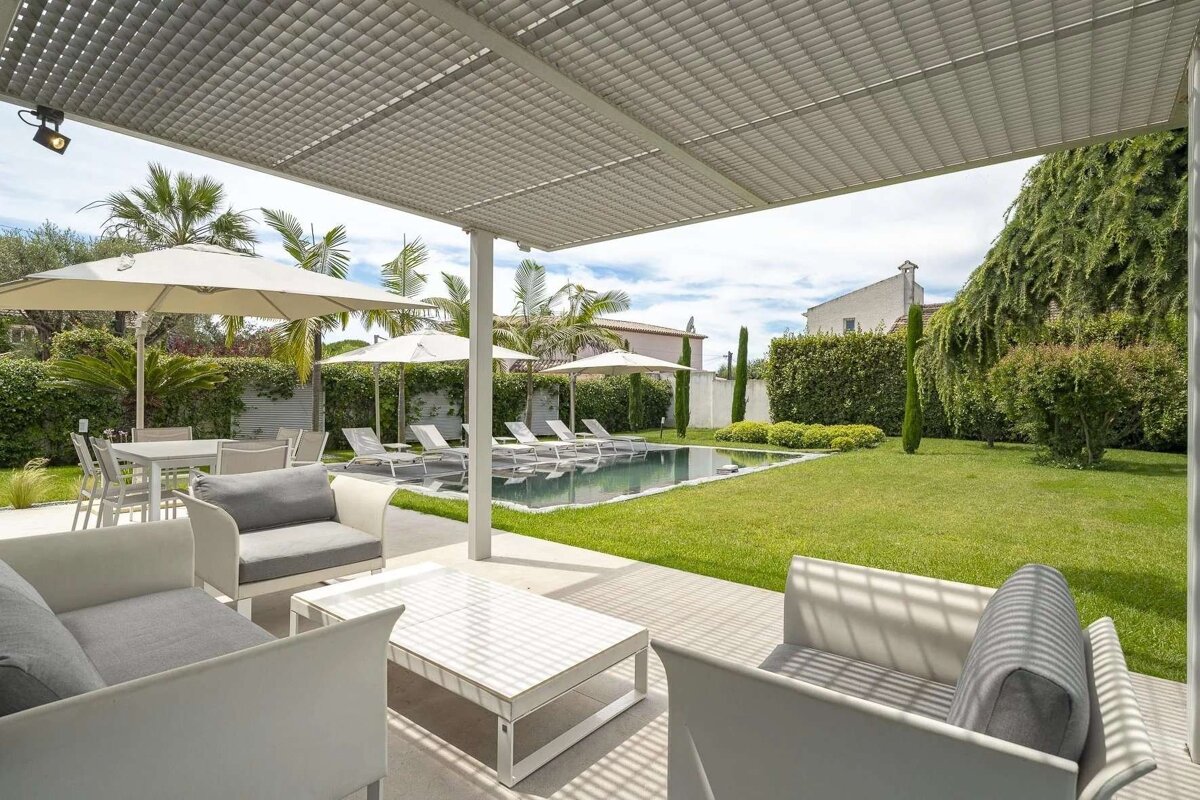 A modern outdoor patio with white lounge furniture under a pergola, a swimming pool, green lawn, and sun umbrellas.