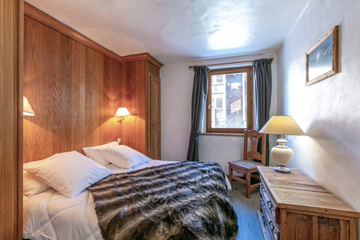 A cozy, rustic bedroom with wood-paneled walls, a bed with a furry throw, a sunlit window, and wooden furniture, exuding a warm, inviting ambiance.