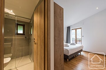 A modern bedroom with a large bed, wooden floor, and balcony is connected to a stylish bathroom featuring a glass shower and toilet.