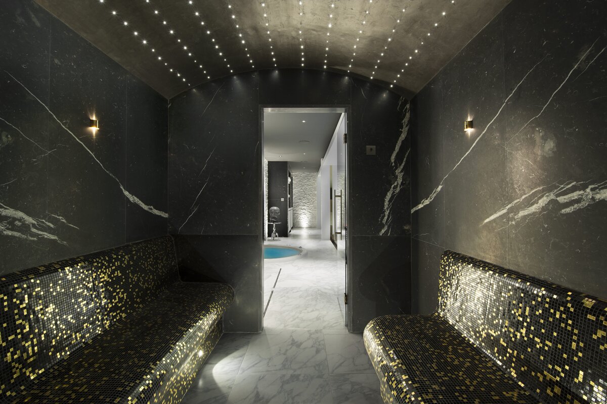 Luxurious spa room with dark marble walls, star-lit arched ceiling, and sparkling black and gold mosaic benches. Beyond is a small pool.