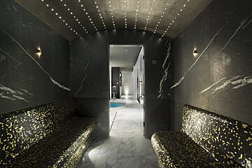 Luxurious spa with dark marble walls, starry ceiling lights, and two gold mosaic benches. A doorway leads to a pool area.