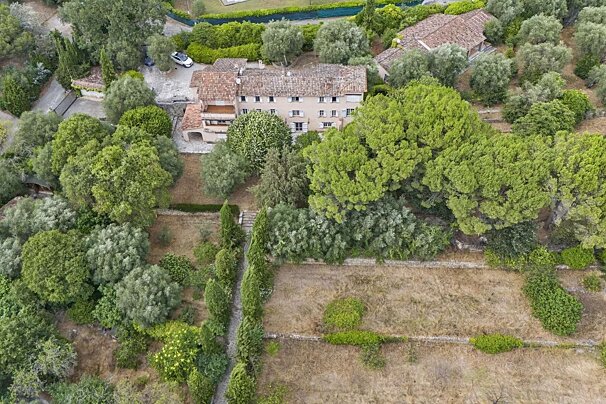 Aerial view of a beige house with a red-tiled roof, surrounded by lush green trees, terraced gardens, and dry grass.