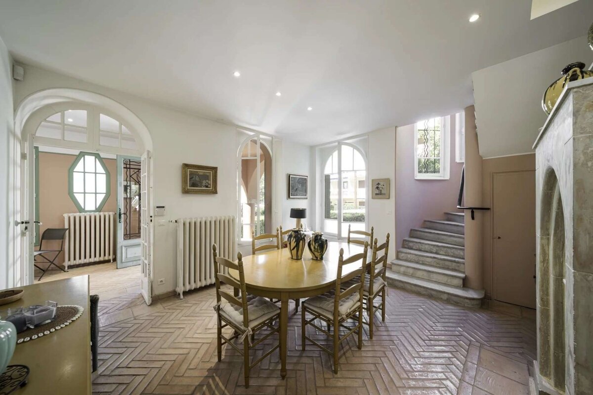 A light-filled dining room features a herringbone tile floor, oval table with chairs, arched windows/doorways, and a winding staircase.