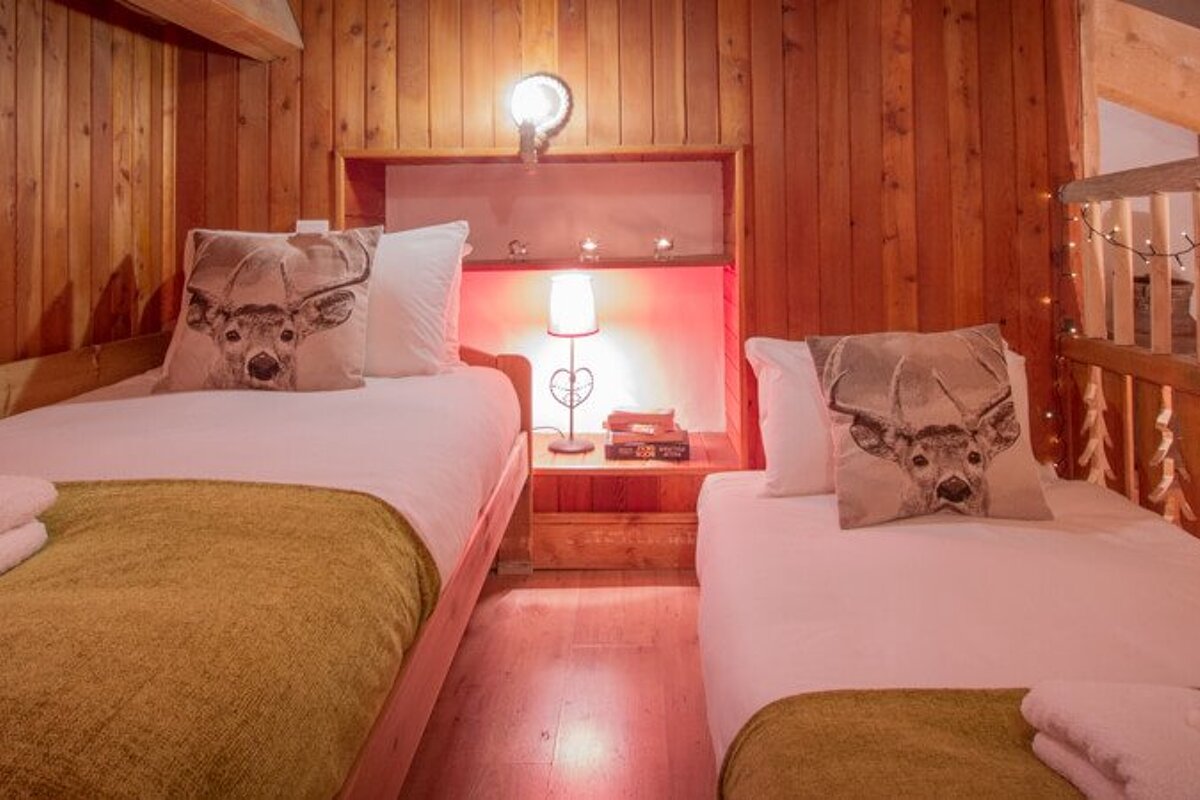 Cozy wood-paneled bedroom in a chalet with two single beds featuring deer-print pillows and a bedside lamp.