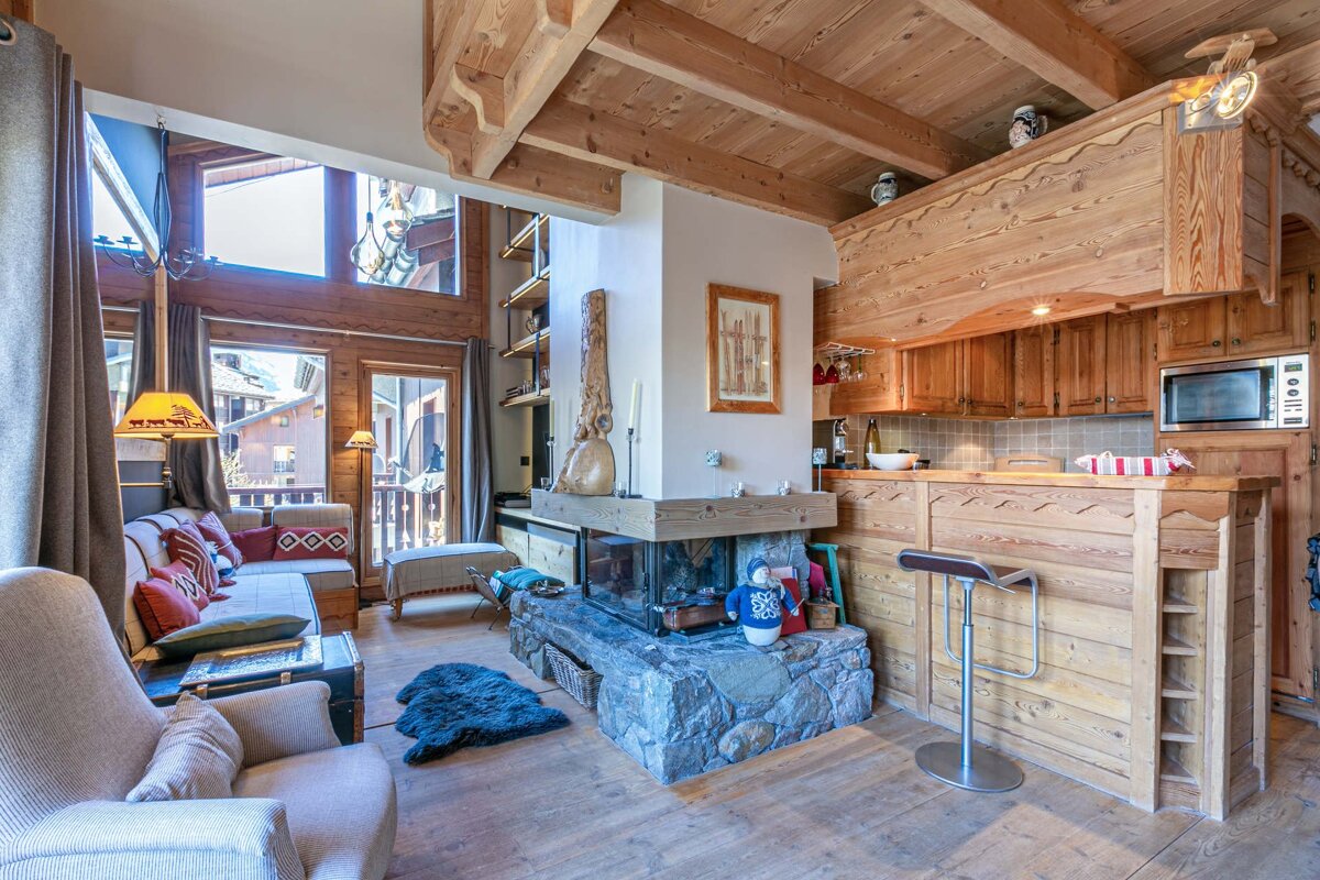 A cozy alpine chalet interior featuring a central stone fireplace, a rustic wooden kitchen with a bar, and comfortable seating.