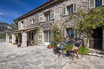 Sunny stone villa with green shutters and climbing vines, overlooking a large paved patio with outdoor seating and distant mountains.