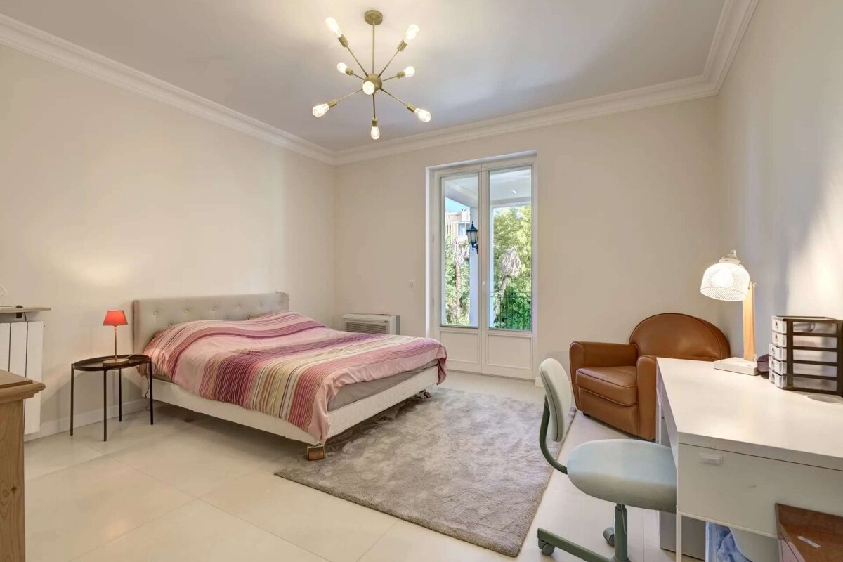 A modern bedroom with a bed, desk, armchair, and French doors. A sputnik chandelier illuminates the light-colored space.