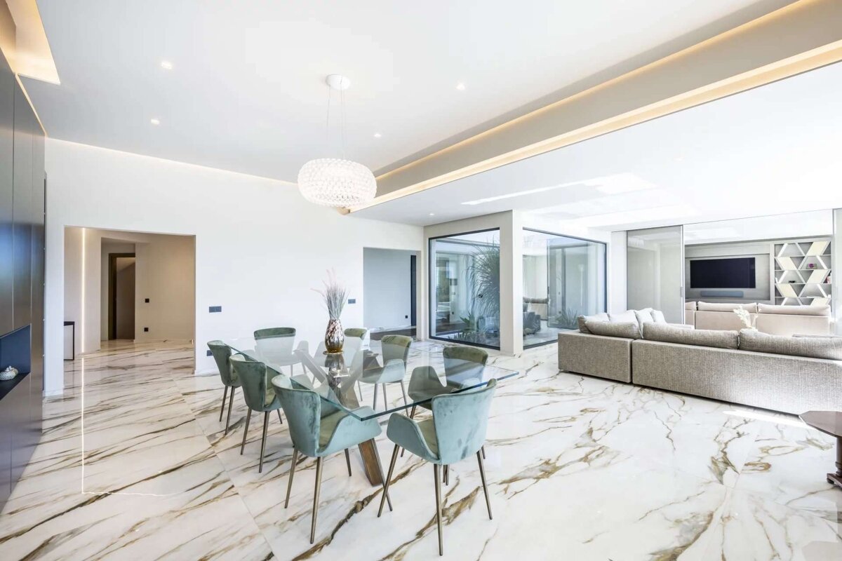 Spacious, modern open-plan living & dining area. Features white marble floors, glass dining table with green chairs, grey sofa, and large windows.