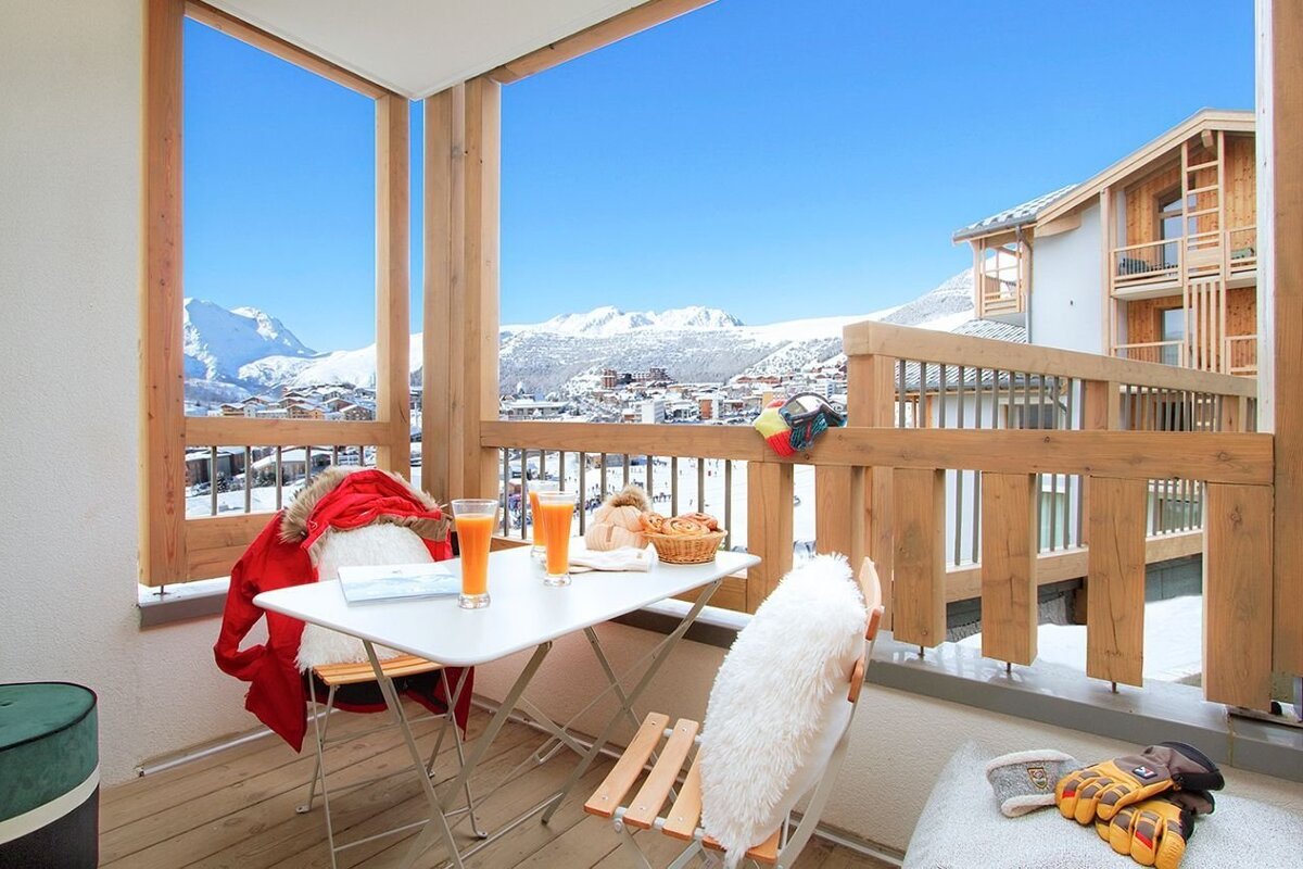 A balcony with breakfast (juice, pastries) and ski gear overlooks a stunning panoramic view of a snowy mountain ski resort under a clear blue sky.
