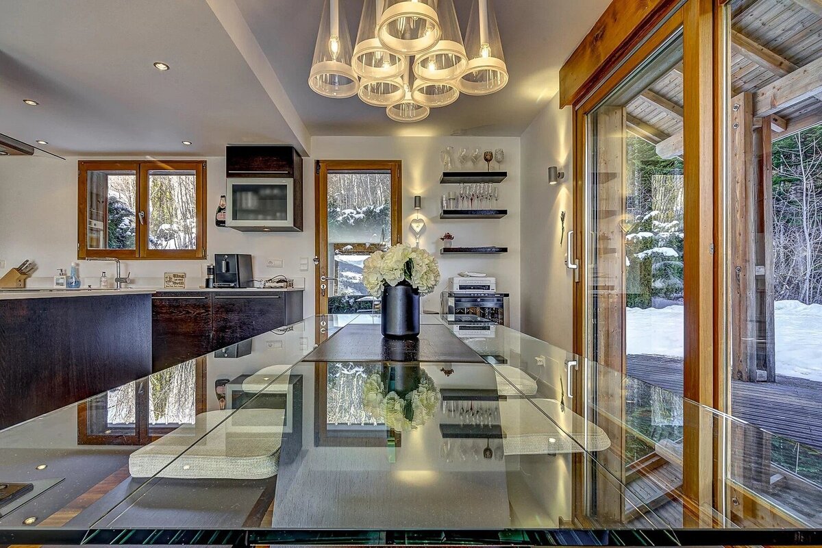 A modern dining area features a sleek glass table, contemporary lighting, and large windows overlooking a snowy outdoor landscape.