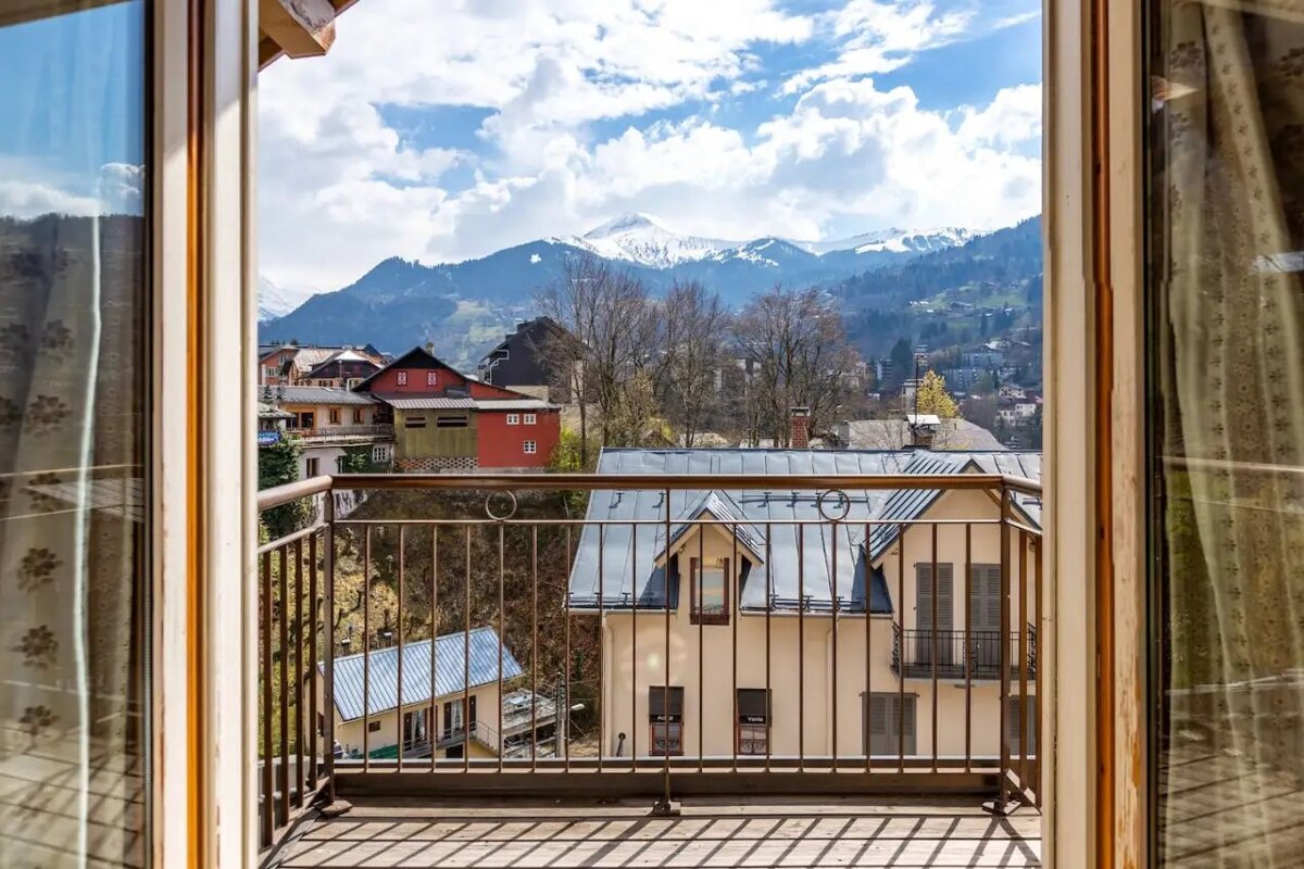 View from a wooden balcony overlooking a scenic mountain village and distant snow-capped peaks under a cloudy blue sky.