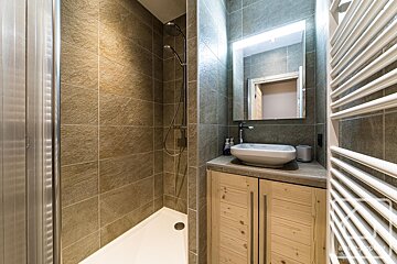 A modern bathroom with a tiled shower stall next to a vanity with a white vessel sink, wooden cabinet, and lit mirror.