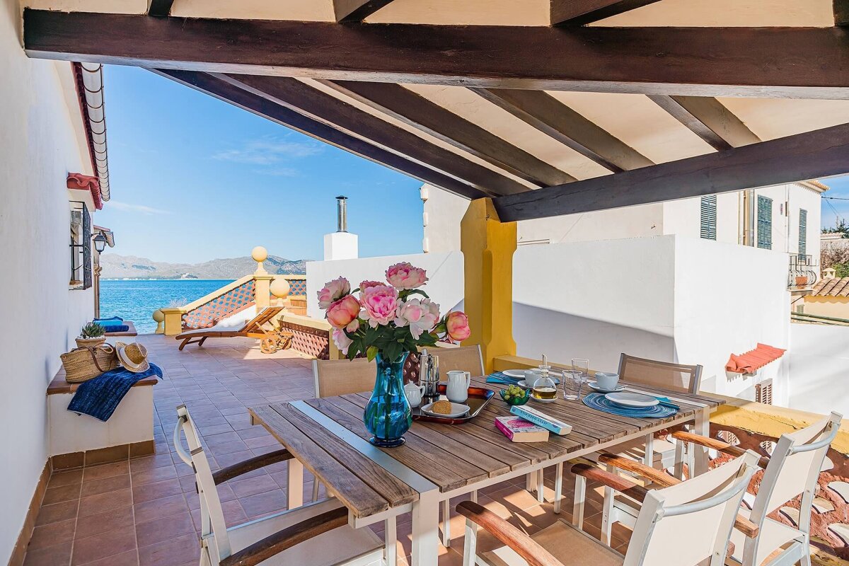 A sunny outdoor terrace with a shaded dining table, set with flowers and food, offers stunning views of a sparkling blue sea and distant mountains.